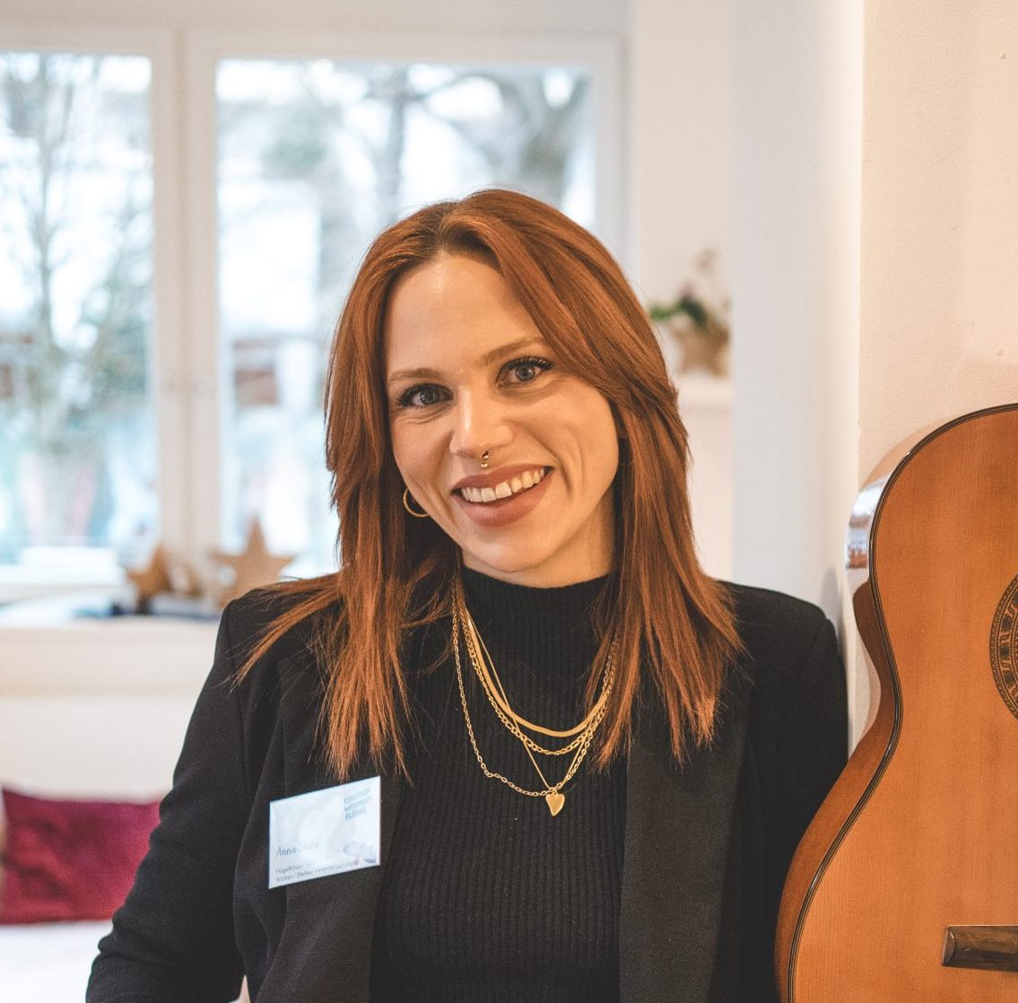 Portr&auml;tfoto von Anna, l&auml;chelnd im schwarzen Blazer neben einer Gitarre