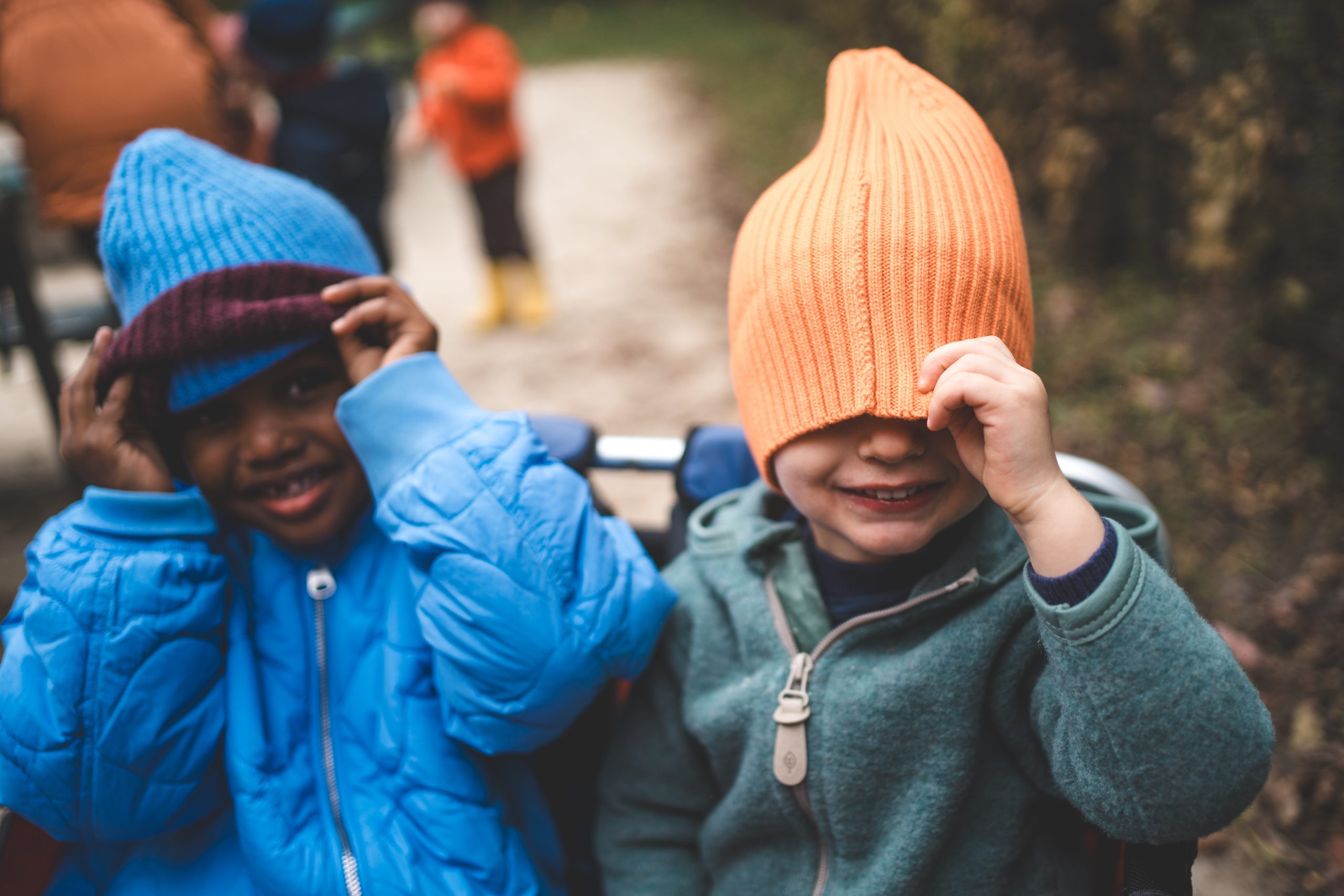 Zwei Kinder mit bunten M&uuml;tzen drau&szlig;en: eines zieht sich lachend die orange M&uuml;tze &uuml;bers Gesicht