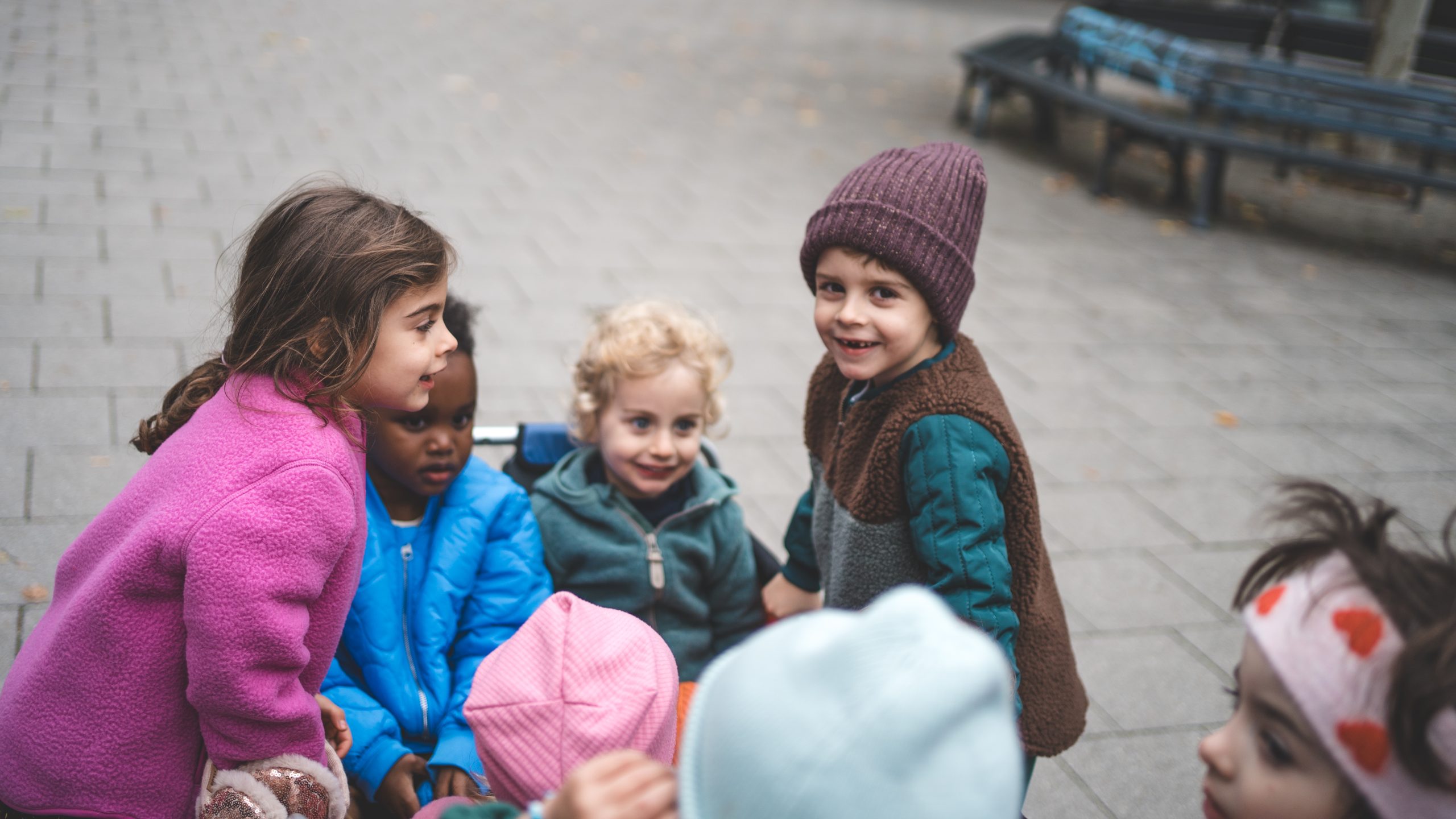 Kinder in bunten Jacken und M&uuml;tzen stehen lachend zusammen im gepflasterten Au&szlig;enbereich der Kita