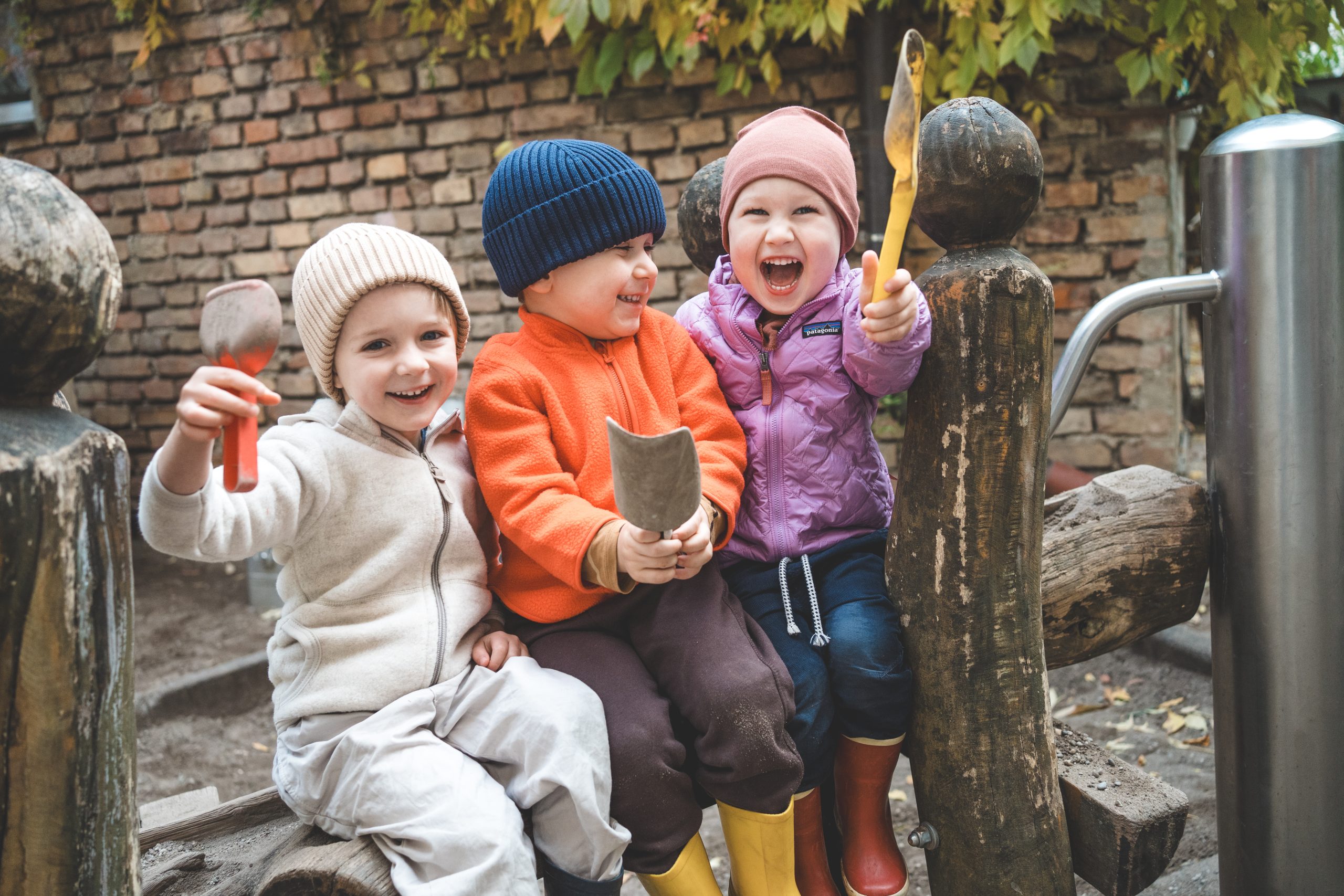 Drei lachende Kleinkinder mit M&uuml;tzen sitzen drau&szlig;en auf einem Holzspielger&auml;t und halten Holzst&uuml;cke in den H&auml;nden