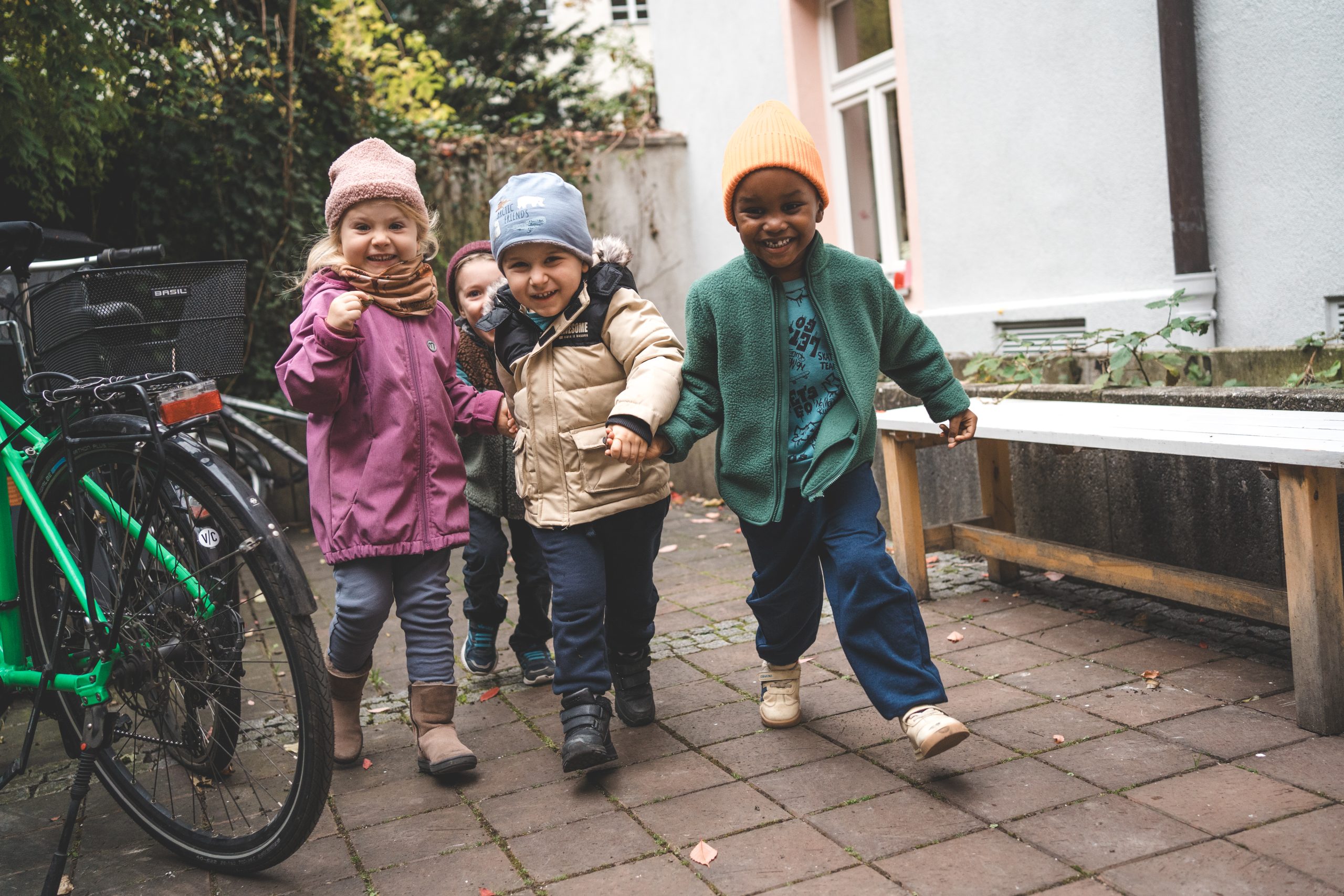 Drei Kinder in Jacken und M&uuml;tzen laufen lachend &uuml;ber den Hof der Kita, daneben ein Fahrrad