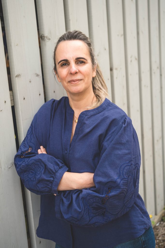 Portr&auml;tfoto von Fernanda in blauer Bluse mit verschr&auml;nkten Armen vor einer hellen Holzwand