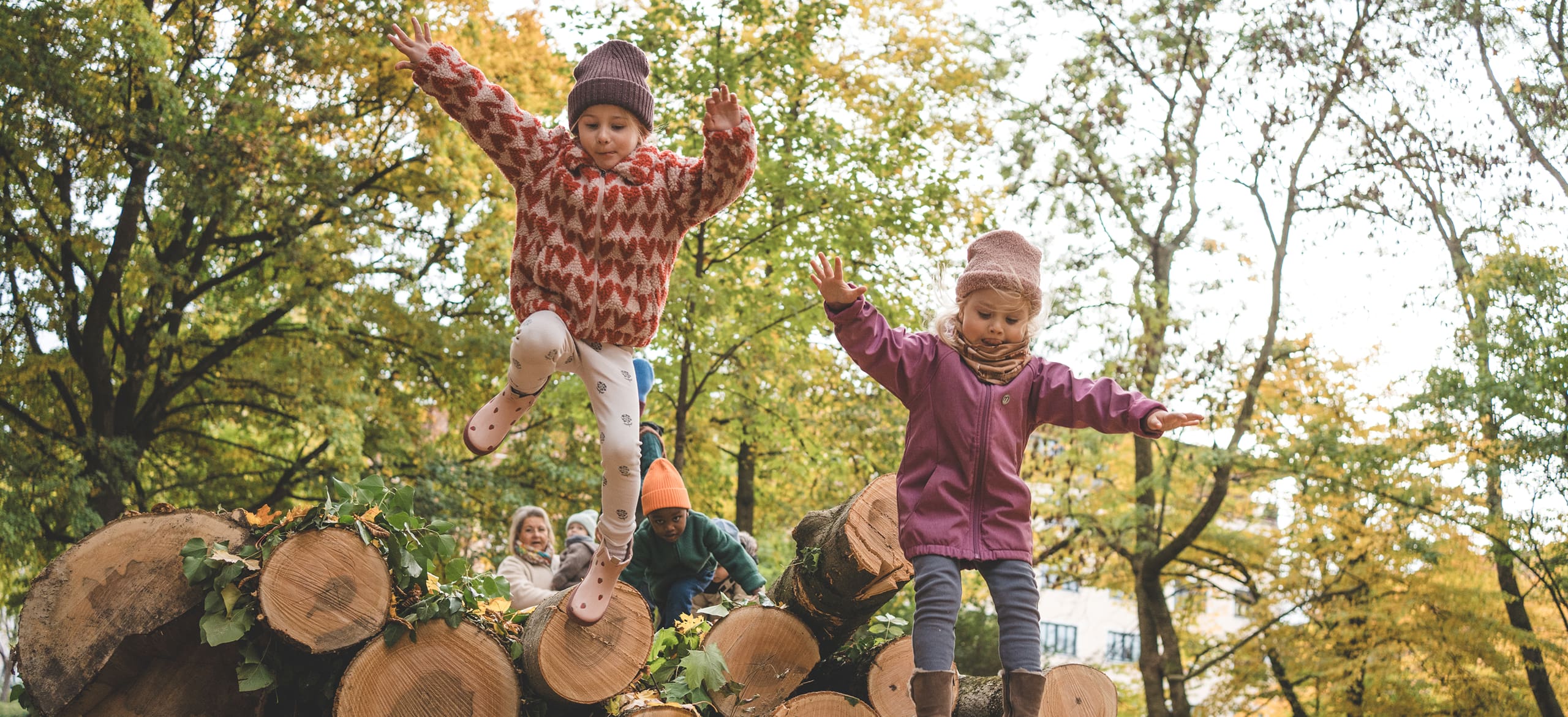 Zwei Kinder springen fröhlich von einem Holzstapel im herbstlichen Wald