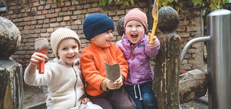 Drei lachende Kleinkinder mit Mützen sitzen draußen auf Holzstämmen und halten Werkzeuge in den Händen